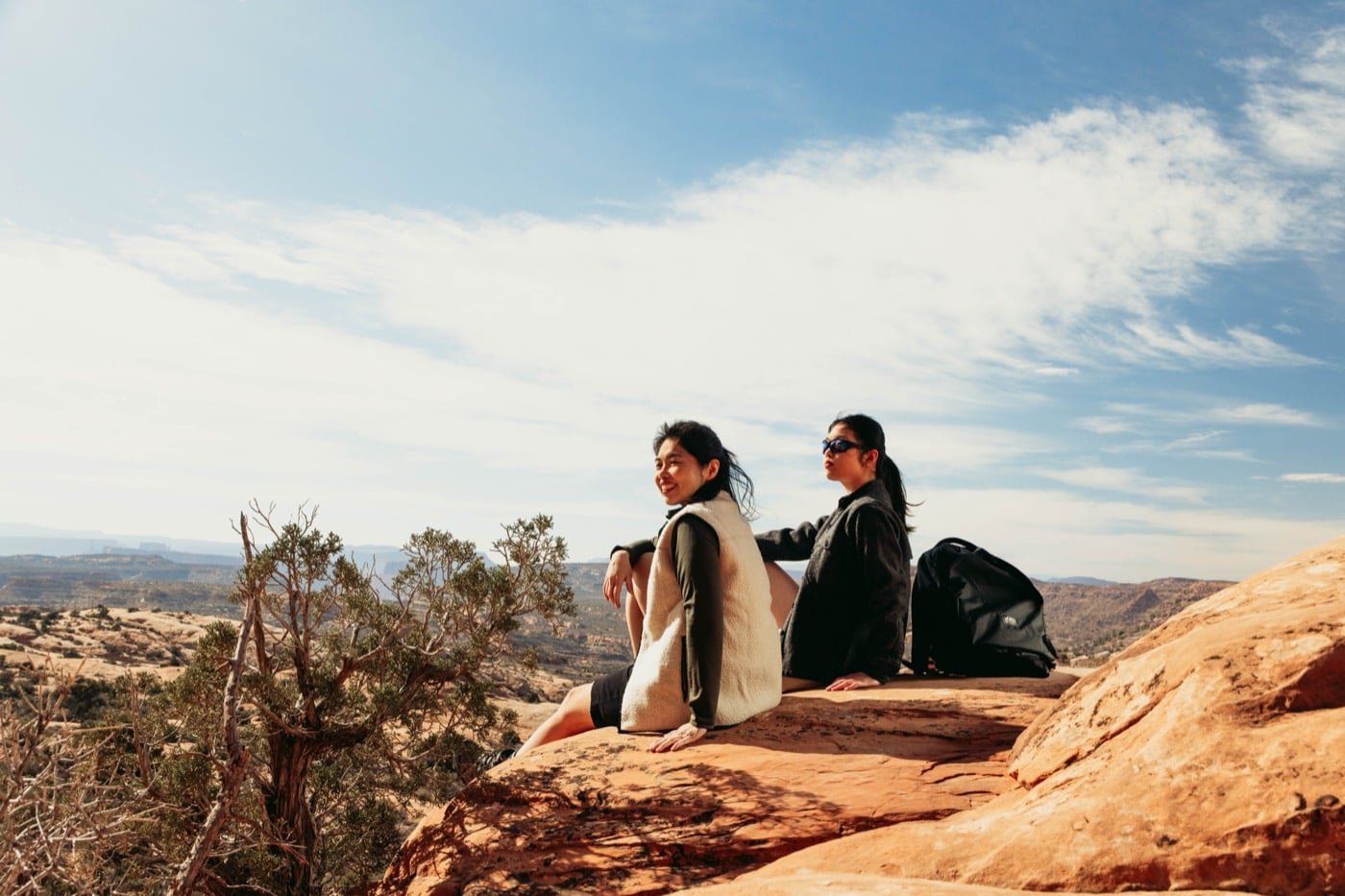 Two friends resting on a sandstone ledge above a canyon in Moab, looking out at the view.